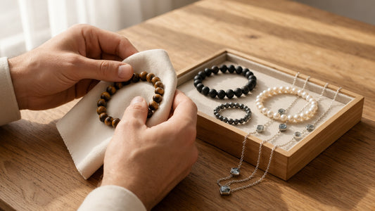 Hände polieren ein Tigerauge-Armband mit einem weichen Tuch, daneben eine Schmuckschale mit Onyx-Armband, Hämatit-Ring, Perlenarmband und Kette